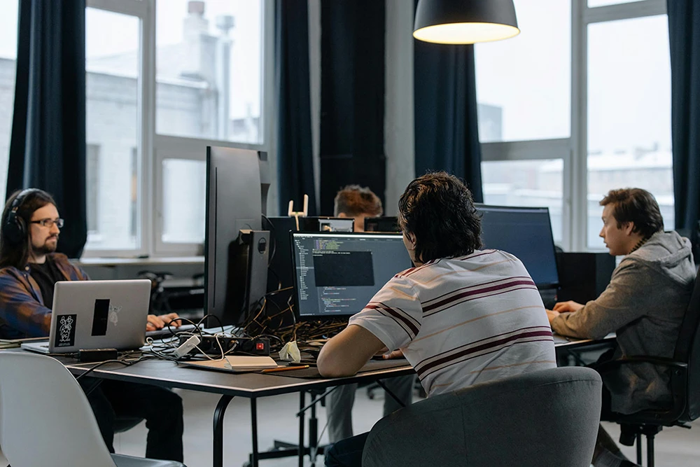 Three IT professionals working at computers in modern office space with dark curtains and natural lighting.