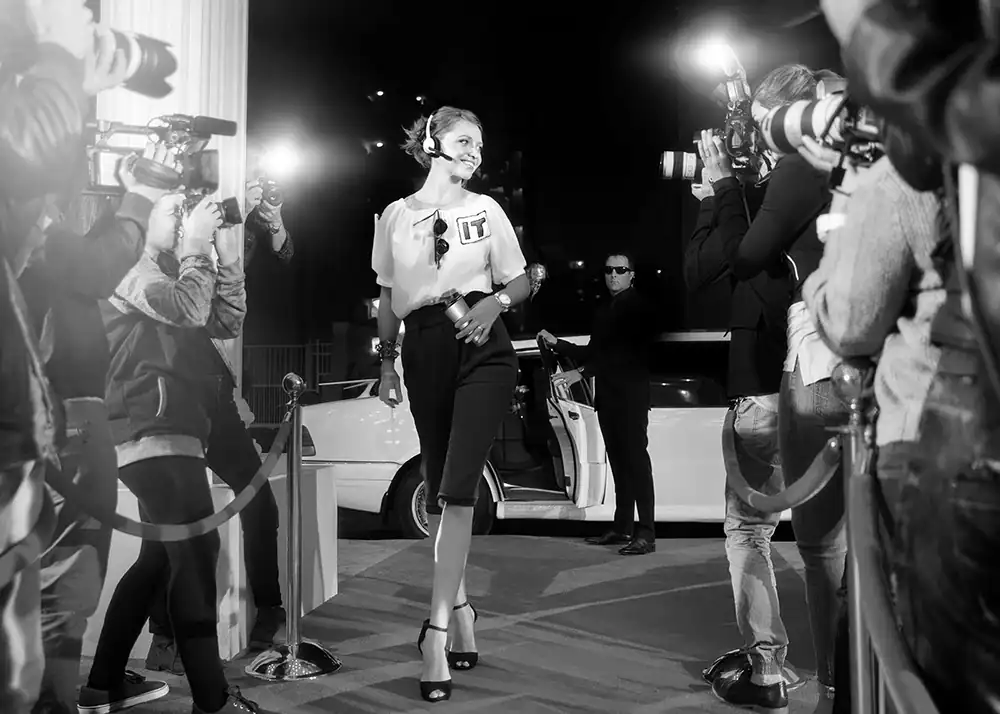 Stylized image of a glamorous woman wearing a headset and a shirt labeled “IT,” walking a red carpet as paparazzi take photos—representing personalized, high-priority tech support.