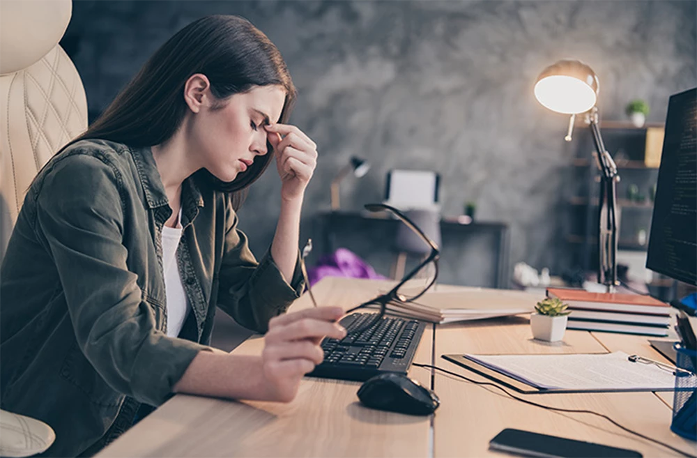 A stressed woman at a desk, holding her glasses and pinching her nose, looking at a computer monitor.