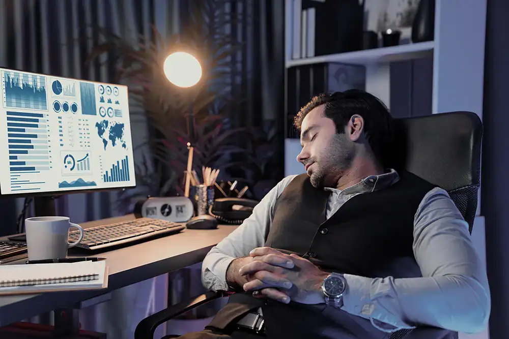 Office worker asleep at desk in front of data charts, symbolizing burnout and delays caused by reduced tech support staff.