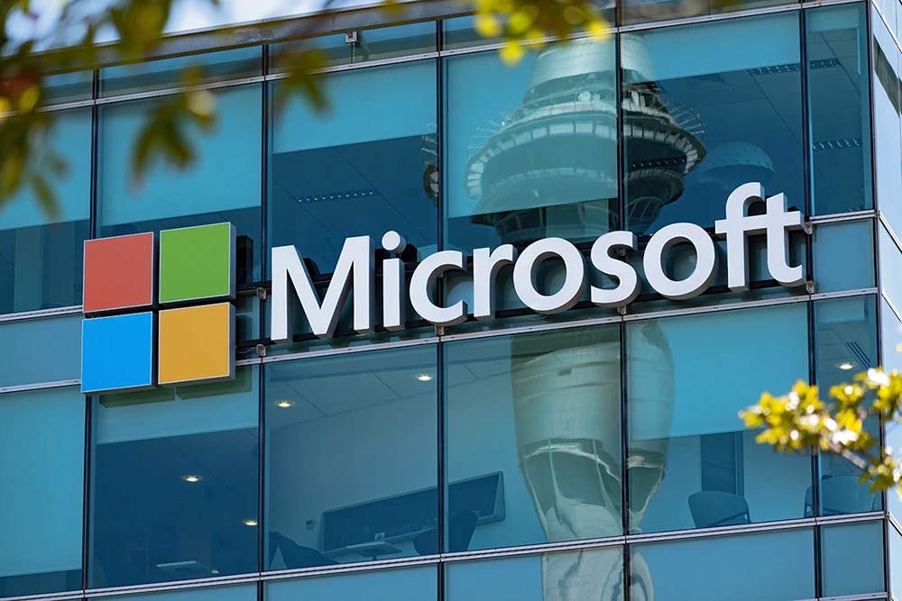 Microsoft logo and corporate branding on a modern glass office building exterior, with a tower reflected in the windows and yellow foliage visible in the foreground.