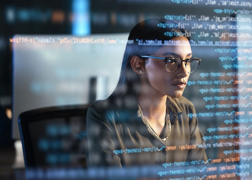 Woman in glasses working at a computer with code reflected on the screen.